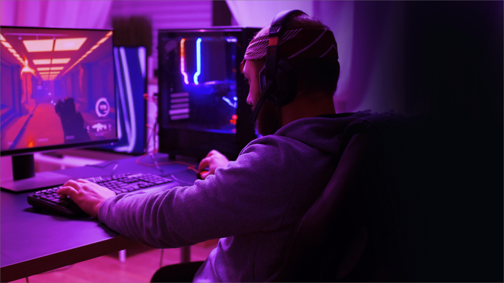 Un joueur assis à son bureau, portant un casque, joue à un jeu vidéo sur un écran d'ordinateur. La scène est éclairée par une lumière violette, avec un PC de jeu lumineux en arrière-plan.